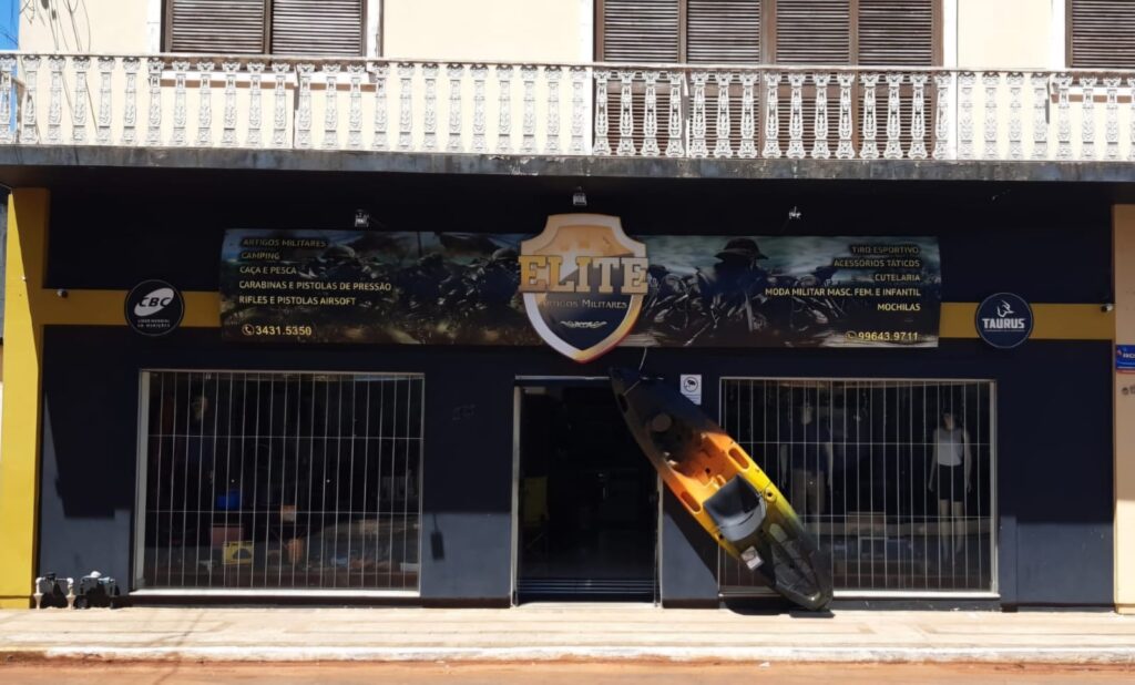 Fachada da loja Elite Artigos Militares em São Borja, RS, especializada em artigos militares, camping, caça e pesca, com carabinas, pistolas de pressão, rifles, pistolas airsoft e moda militar masculina, feminina e infantil. O destaque da fachada inclui o logo da loja e produtos expostos, como um caiaque.